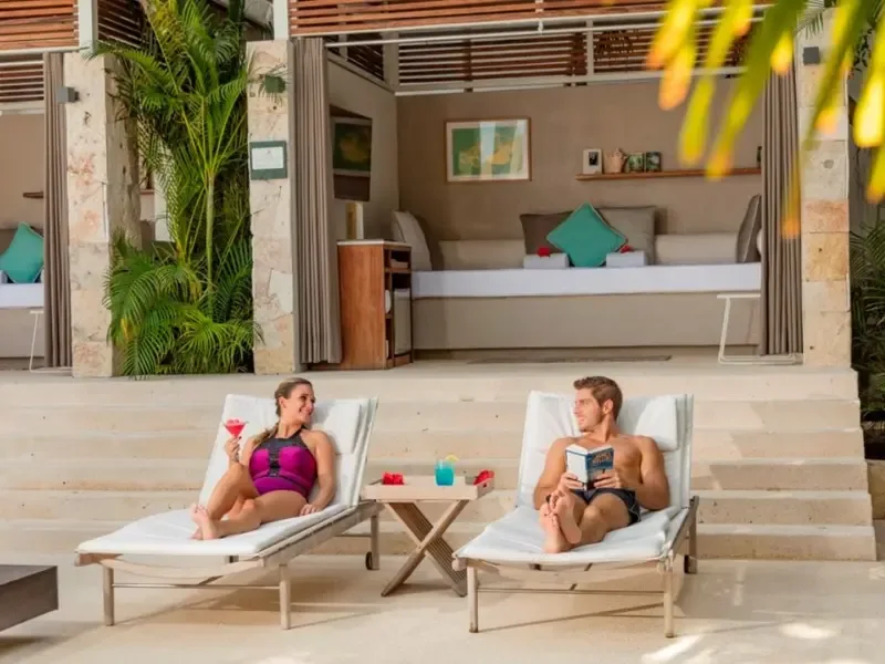 A man and a woman lounging by the pool, enjoying drinks and reading a book in a tropical setting.$# CAPTION