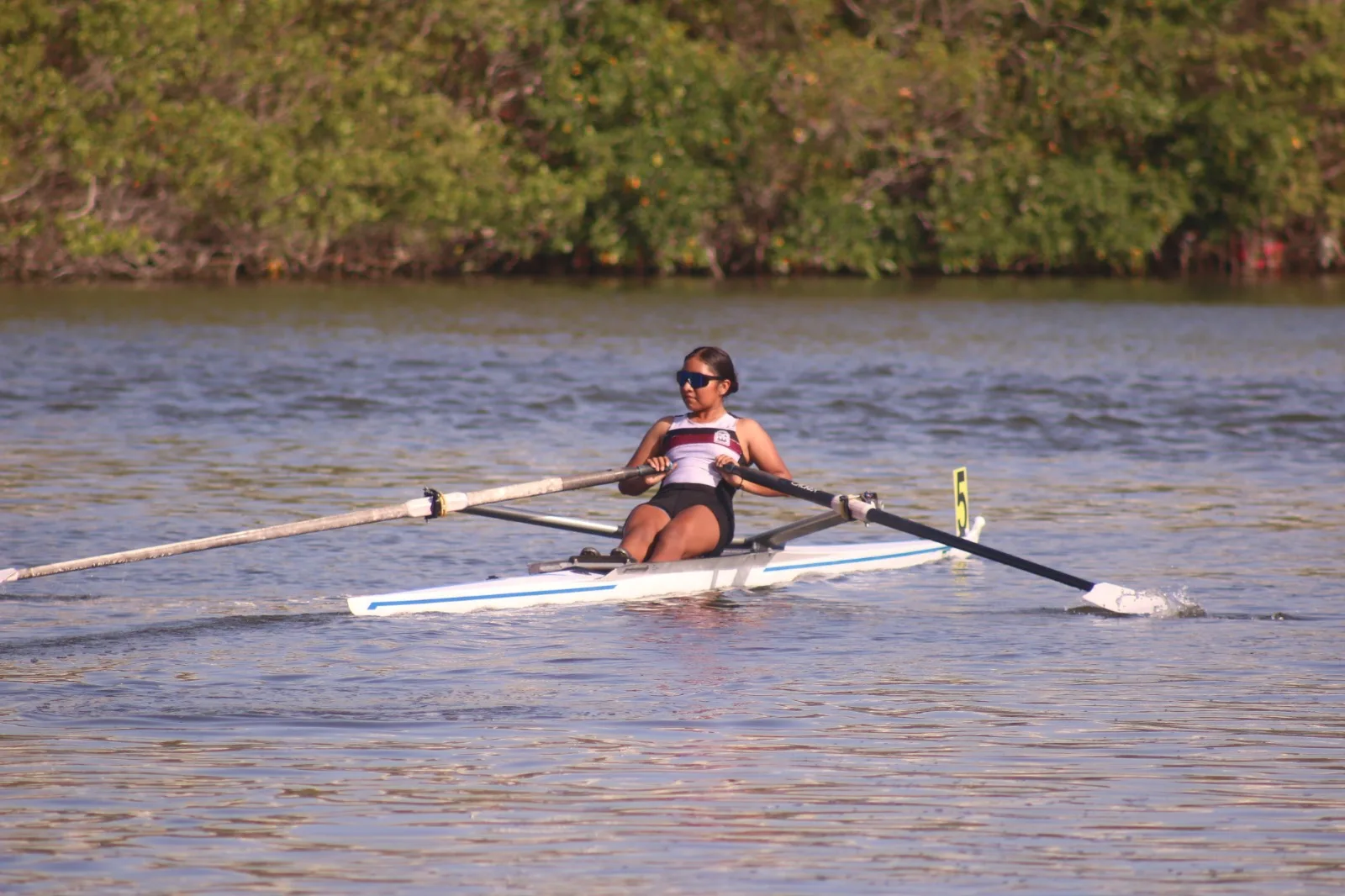 A young woman rowing on calm water surrounded by greenery.$#$ CAPTION