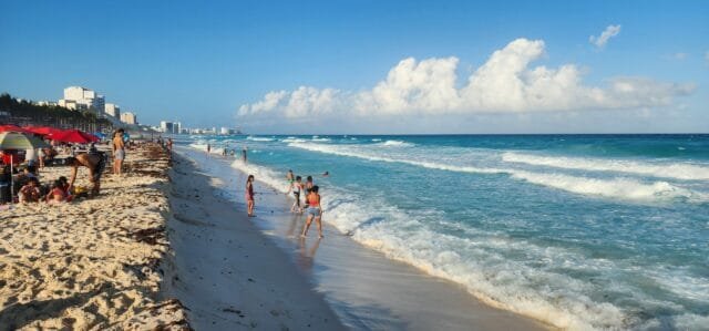 A picturesque beach in Cancun with people playing and relaxing by the water, surrounded by soft waves and a clear sky.$#$ CAPTION