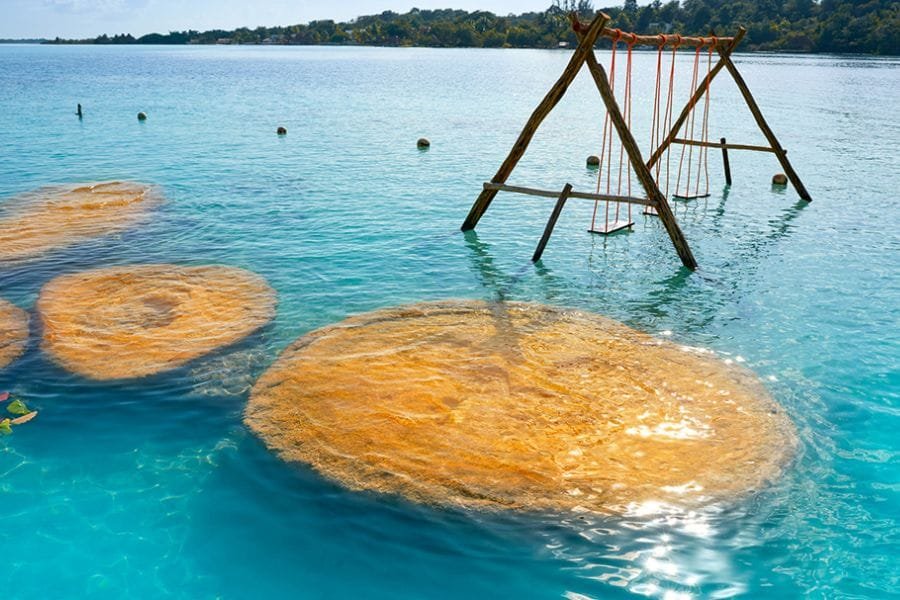 A wooden swing set on a sandy platform in shallow turquoise waters, surrounded by various rock formations.$# CAPTION