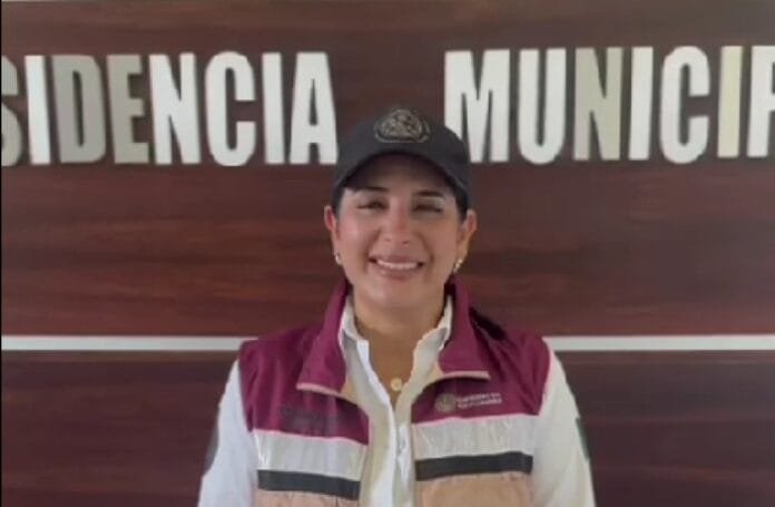A smiling person wearing a cap and a vest stands in front of a wooden wall with the words "PRESIDENCIA MUNICIPAL" visible in the background.$#$ CAPTION