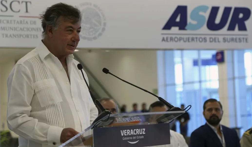 A man in a white shirt delivers a speech at the Veracruz Airport inauguration ceremony, with attendees seated behind him and an airport logo visible above.$# CAPTION