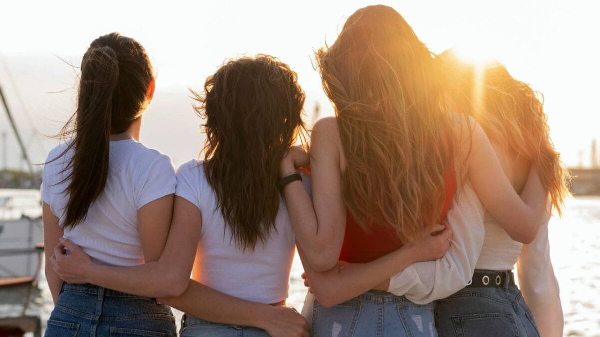 Four friends standing together with their backs to the camera, enjoying a sunset by the water.$# CAPTION