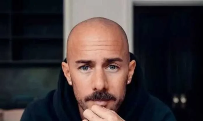 A man with a bald head and a mustache, wearing a black hoodie, looks thoughtfully at the camera with a slight frown.