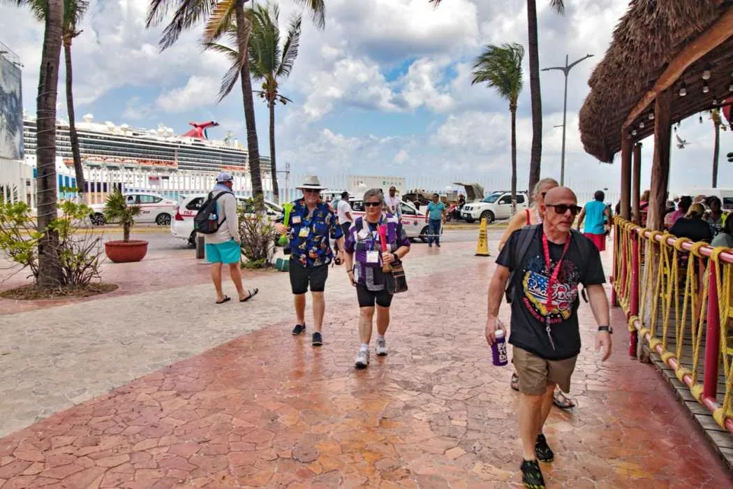 Tourists walking along a pathway near a cruise ship terminal, with palm trees and parked cars in the background
