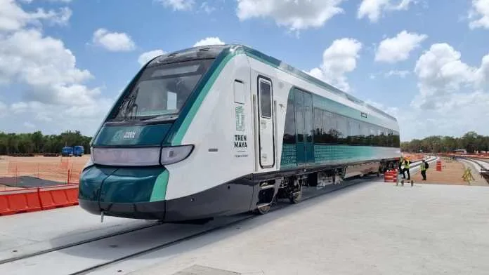 A modern Tren Maya train parked on tracks at a construction site, with workers visible in the background. Clouds are scattered in the blue sky.