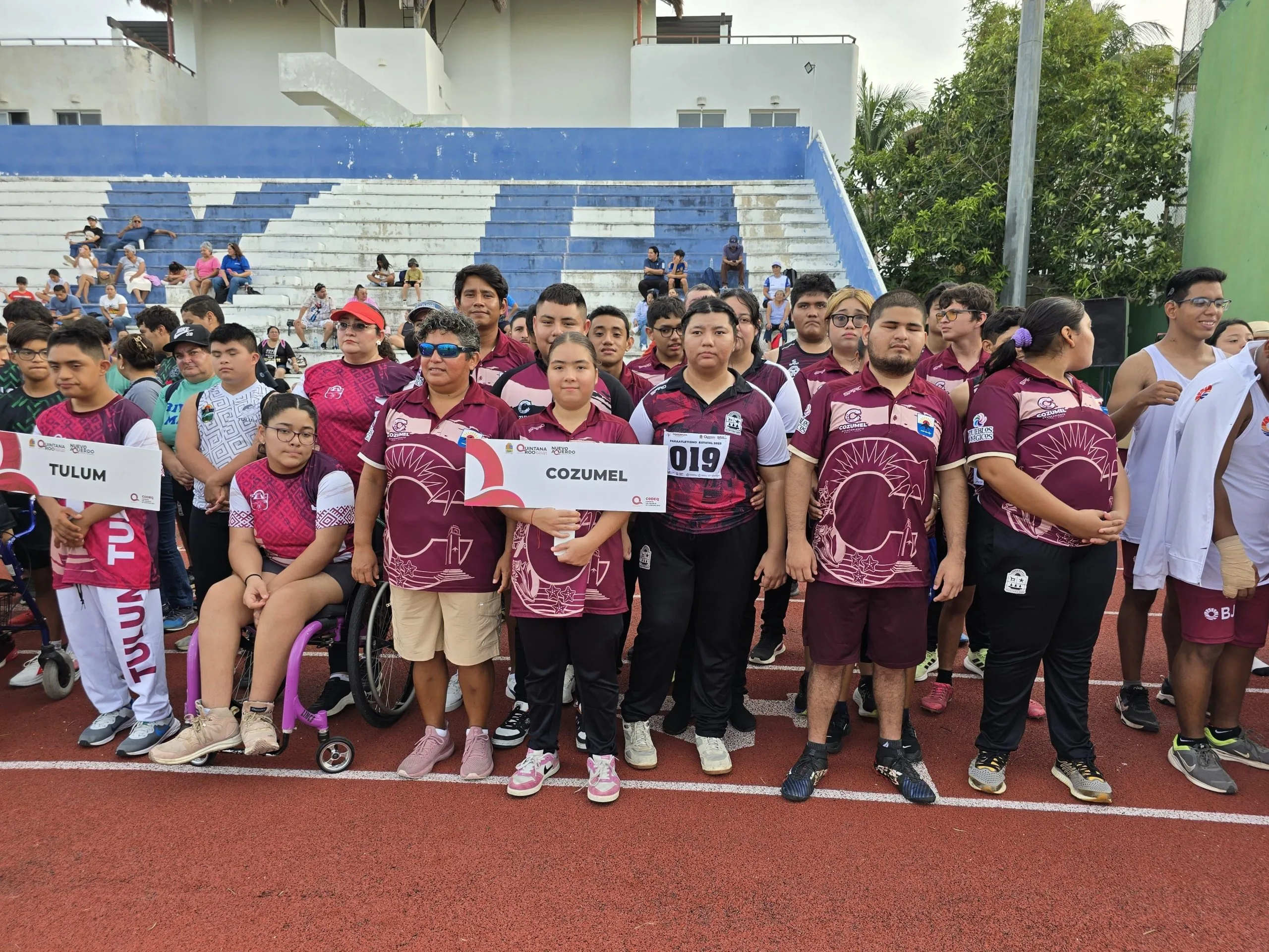 Group of young athletes from Cozumel and Tulum holding signs at a sports event, with spectators in the background.$#$ CAPTION