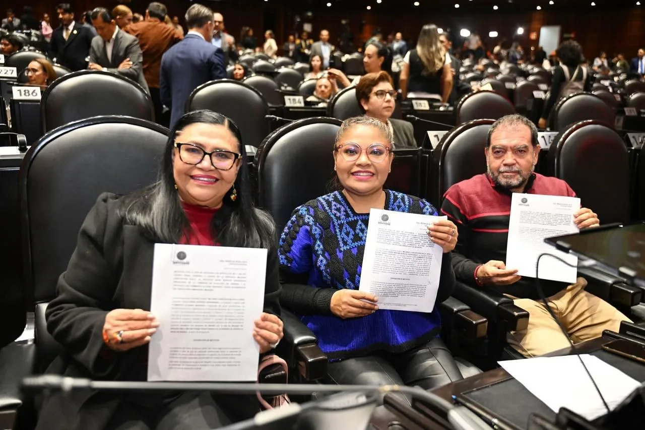 Three individuals holding documents while seated in a legislative assembly, smiling at the camera
