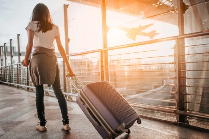 A woman with a suitcase walking through an airport terminal, with an airplane flying outside in the background during sunset