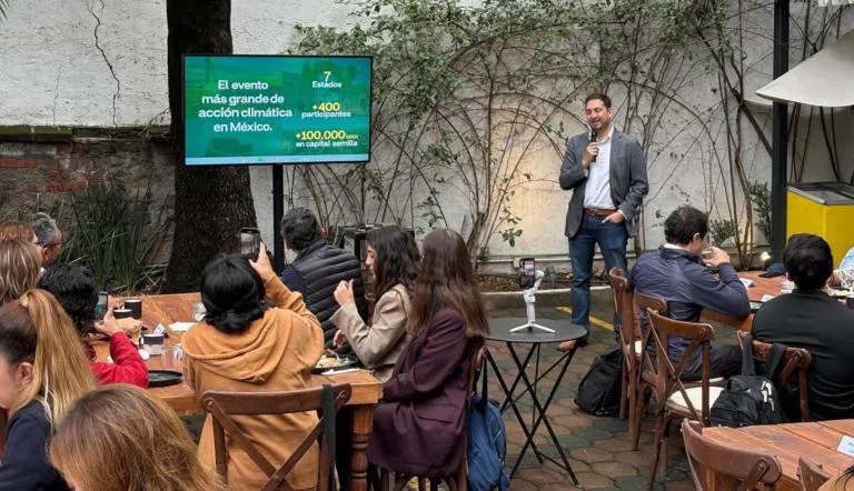 A speaker presenting at a climate action event with a large screen displaying statistics in the background, surrounded by attendees at tables.$# CAPTION