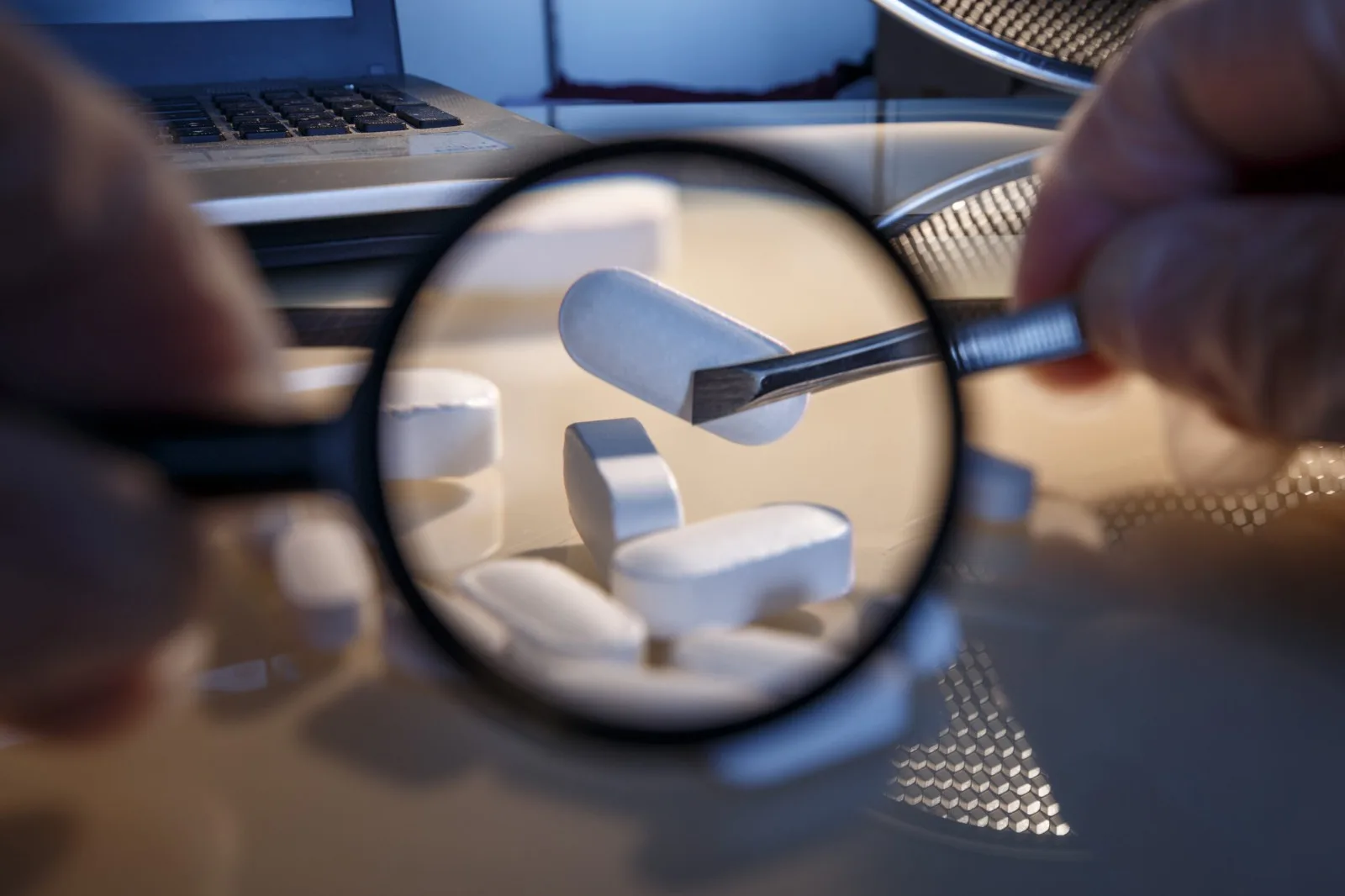 A close-up view of a hand holding a magnifying glass over white tablets with a pair of tweezers poised to pick one up. A laptop is visible in the background.$# CAPTION