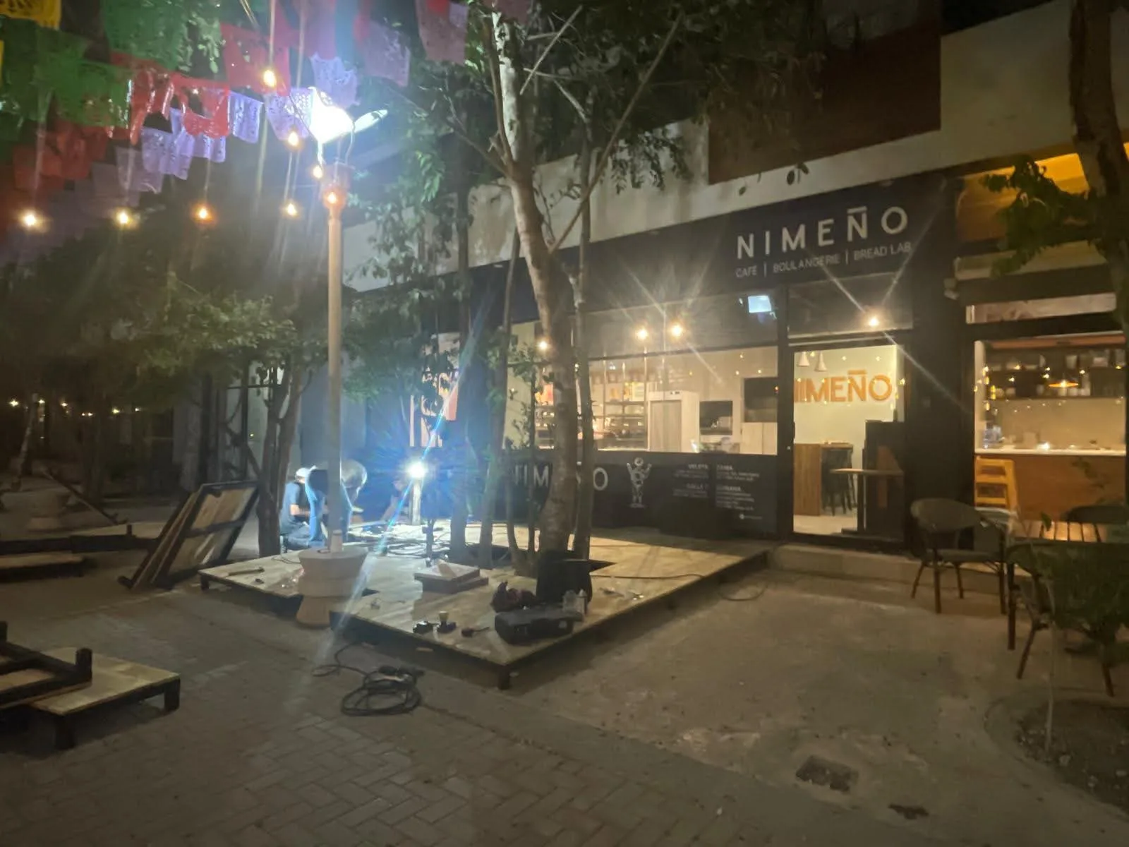 A dimly lit outdoor area of Nimeno Café under construction, with workers and tools visible, surrounded by colorful paper banners and trees in the background.$# CAPTION