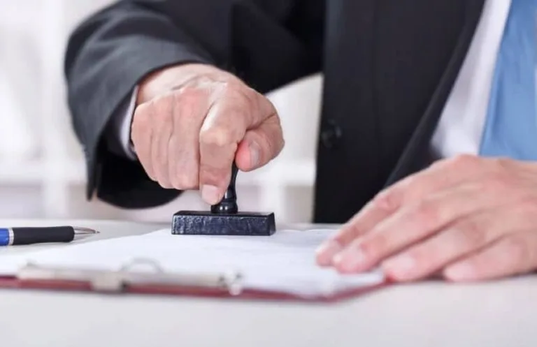 A person's hand pressing a stamp onto a document on a table, wearing a suit with a blue tie. A pen and a clipboard are also visible.