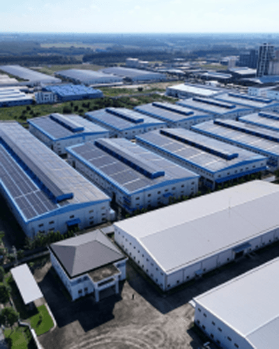 Aerial view of an industrial complex featuring large buildings with solar panels on their roofs, surrounded by green fields and distant structures in the background.$#$ CAPTION