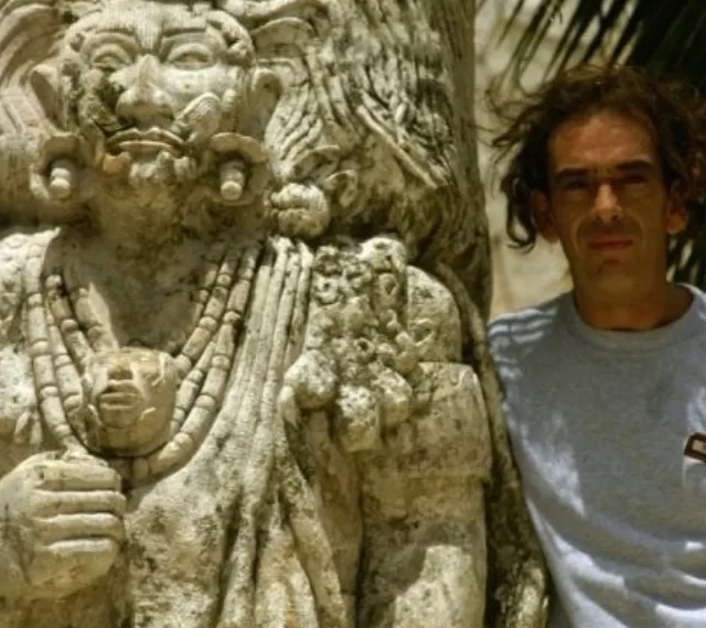 A weathered stone statue of an ancient figure stands next to a man with tousled hair, wearing a gray shirt, in a sunlit environment surrounded by palm leaves.$# CAPTION