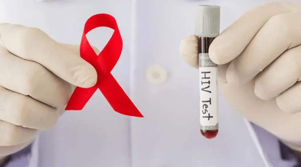 A healthcare professional in gloves holds a red ribbon symbolizing HIV awareness in one hand and a test tube labeled "HIV Test" in the other hand.$# CAPTION