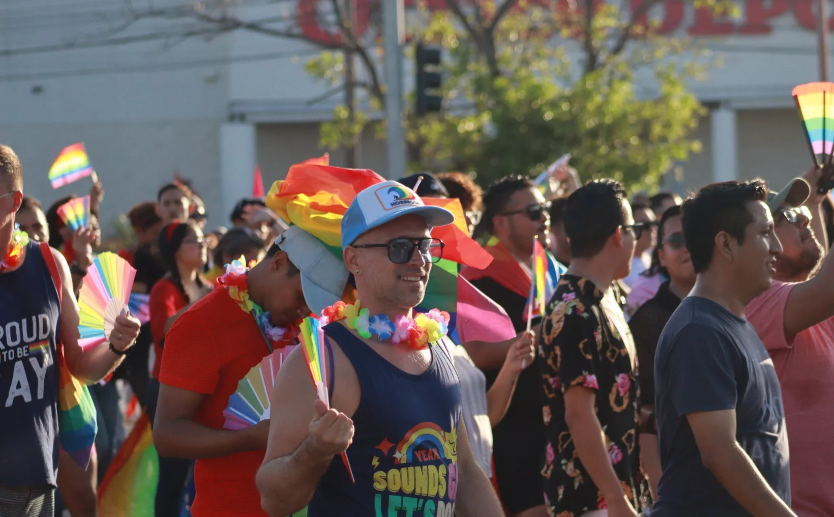 A vibrant pride parade with people holding rainbow flags and wearing colorful attire. A man smiles in the foreground, adorned in a floral lei and a cap, while others participate in the festivities around him.