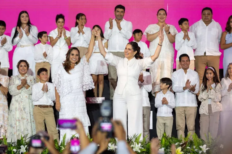 Two women in elegant attire raise their hands together on stage while a group of people applauds behind them. The backdrop features vibrant colors, and the crowd includes children and adults dressed in white.