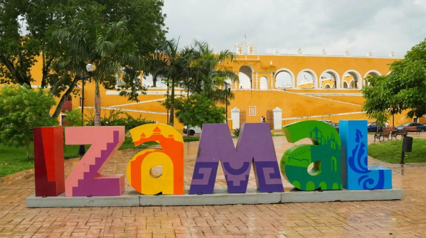 A vibrant sign displaying the name "Izamal" in colorful letters, set against a backdrop of a yellow historic building and greenery-$# CAPTION