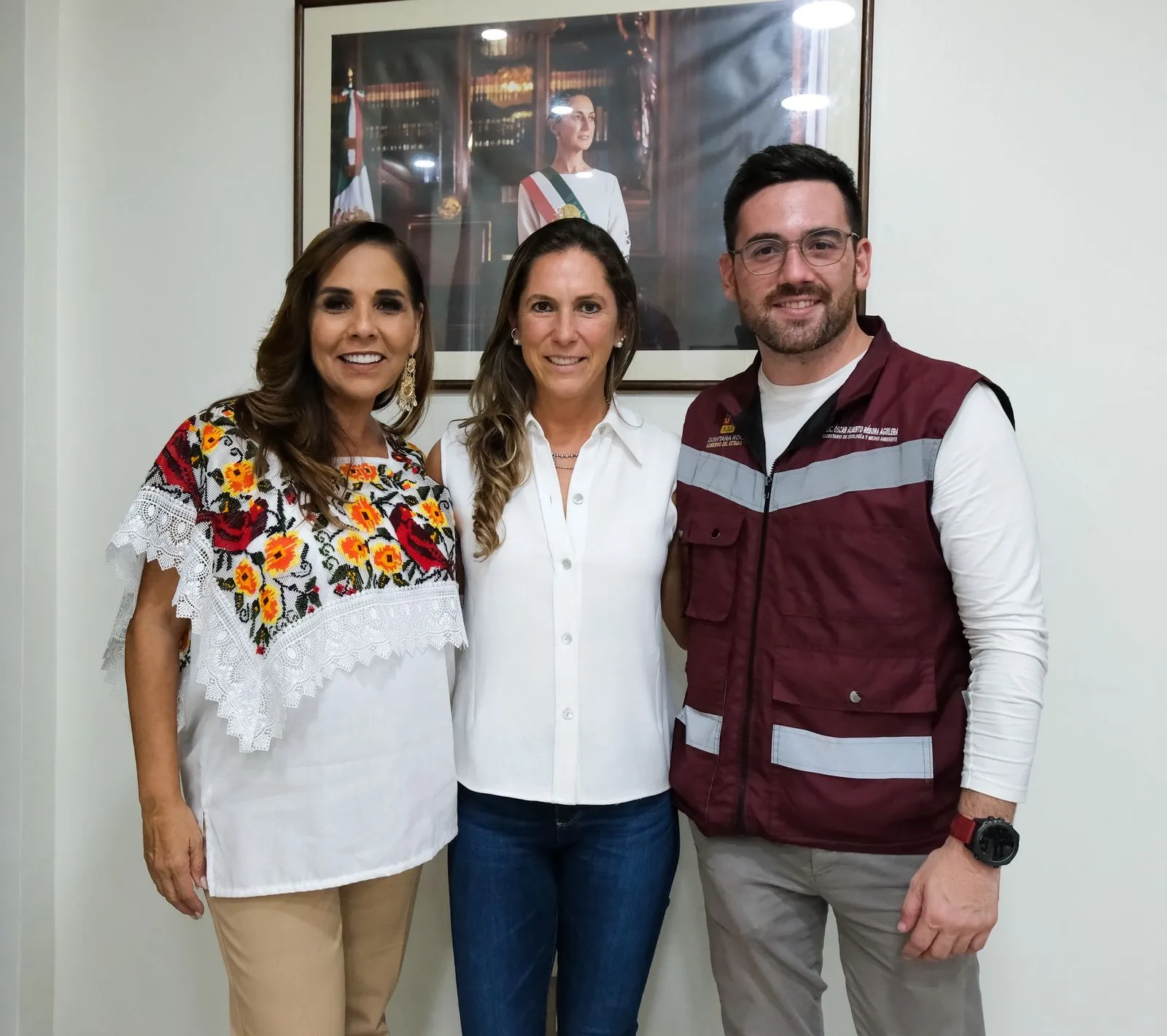 Three individuals posing for a photo against a background featuring a portrait and national flags. One wears a colorful embroidered top, another a white blouse, and the last a red vest with reflective stripes.