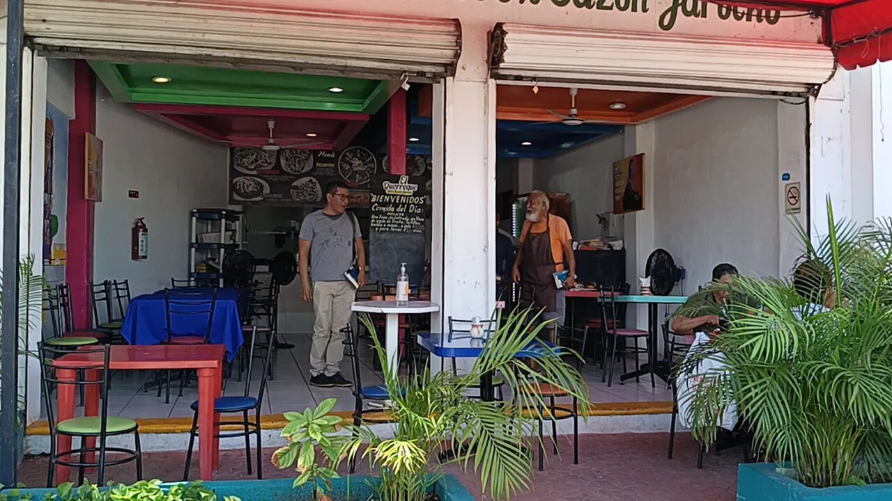 Two men interacting outside a colorful restaurant, with tables and chairs visible, and other diners inside.$# CAPTION