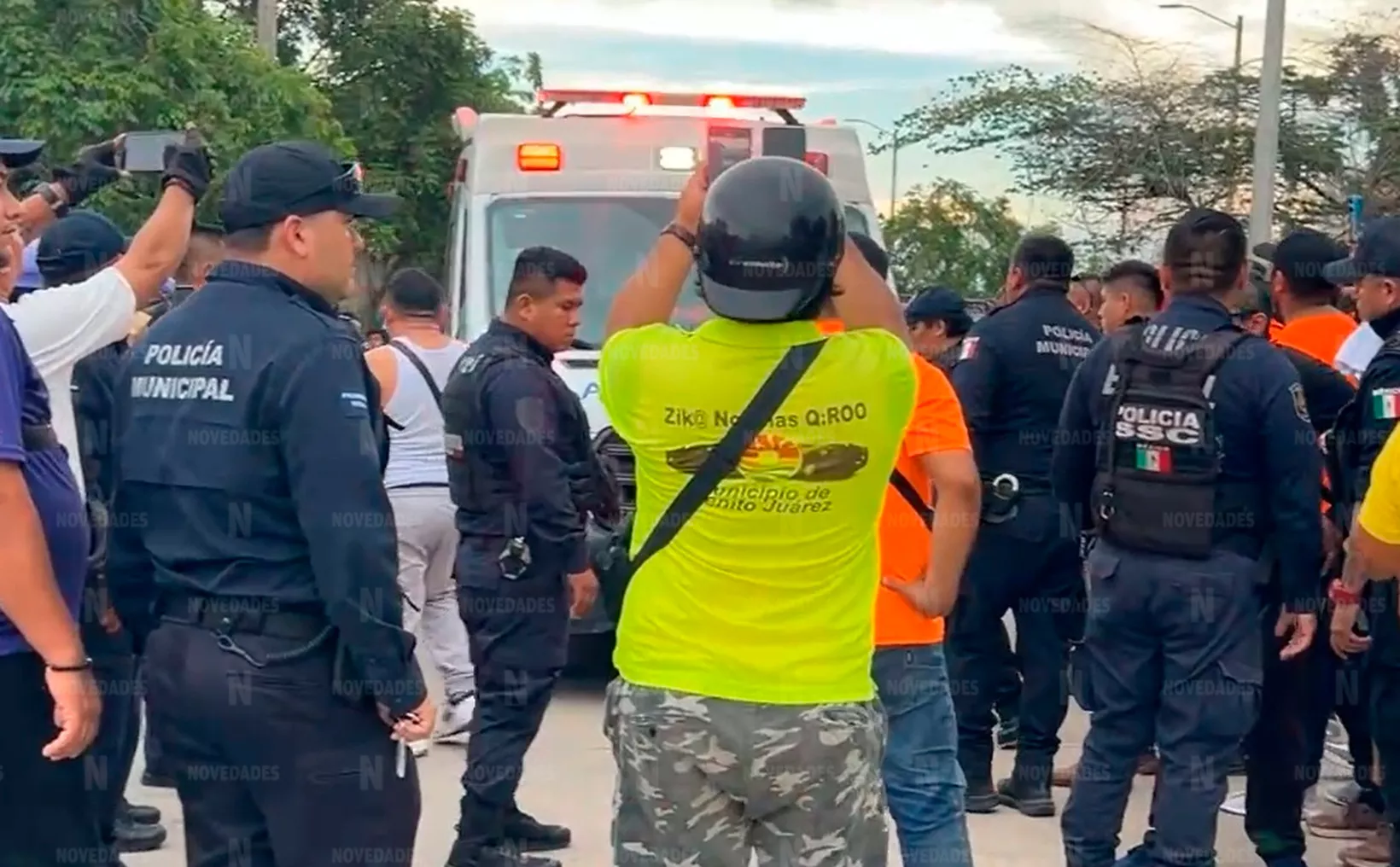 A group of police officers and civilians gathered around an ambulance, with one person taking a photo. The scene appears to be an emergency response situation.