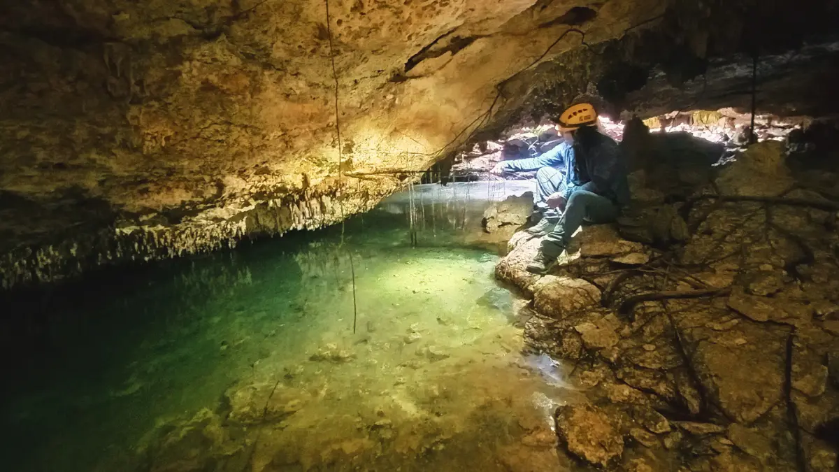 A person in a helmet sits on a rock near a pool of water inside a cave, surrounded by stone formations and dim lighting.$# CAPTION