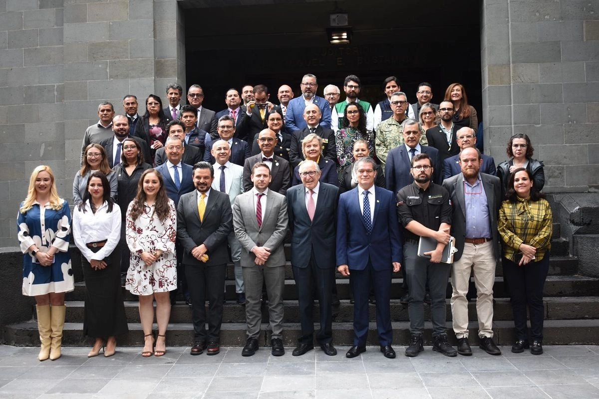 A large group of people, including men and women in formal attire, posing for a photo outside a building. They are standing on steps, with some smiling and others looking serious. The background features a stone wall and an entrance to a building.$# CAPTION