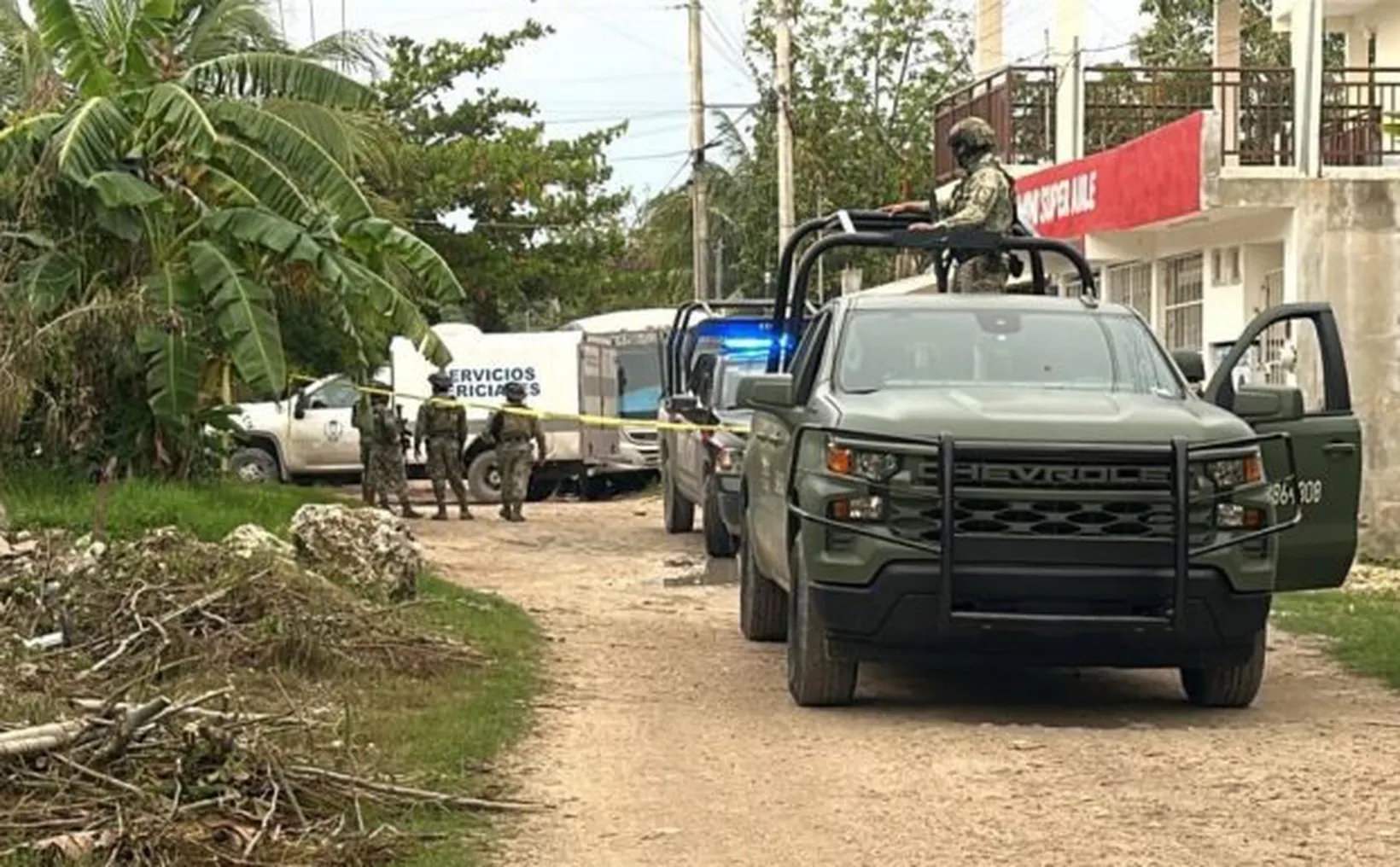A military vehicle is parked on a dirt road, with armed personnel near a service truck and tropical vegetation surrounding the area. Blue lights are flashing from one of the vehicles.