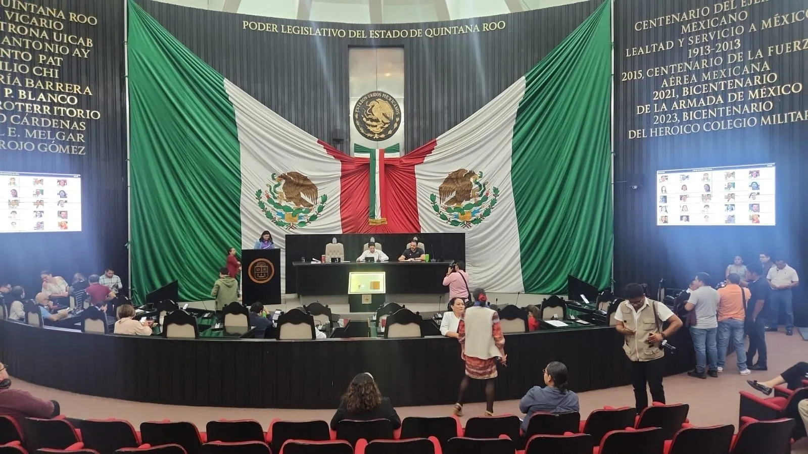 A view of the Quintana Roo Legislative Assembly chamber, featuring a large Mexican flag backdrop and various individuals engaged in discussions and activities.$# CAPTION