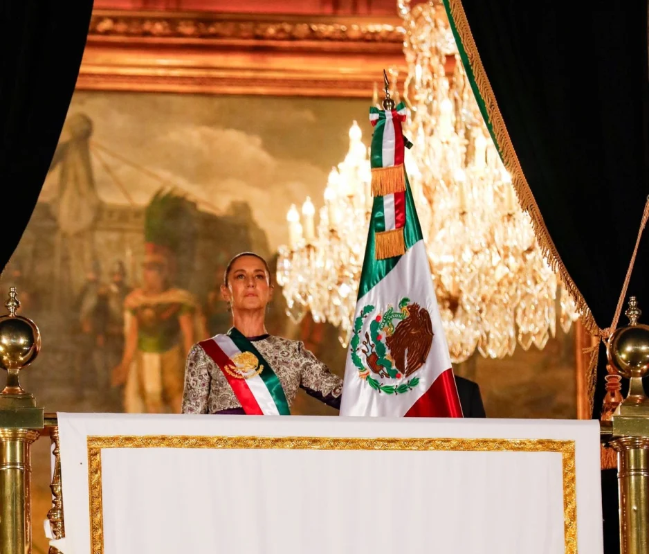A woman stands at a podium, holding the Mexican flag, with a historic painting and chandelier in the background.$# CAPTION