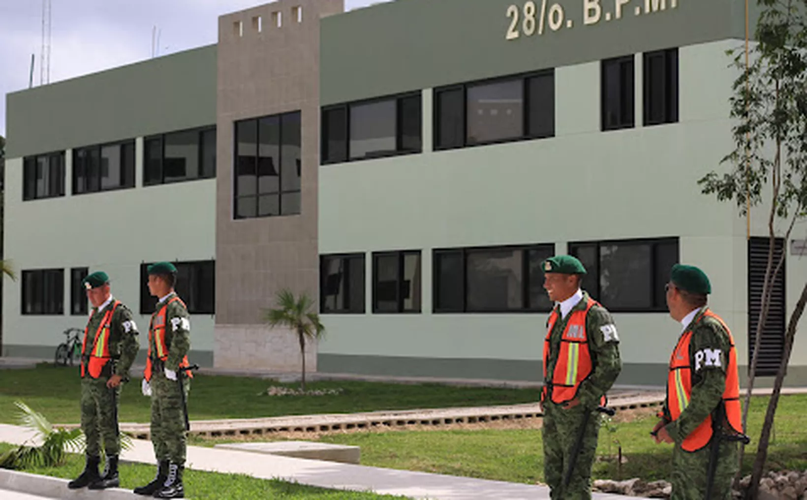 Four soldiers in camouflage uniforms and green berets standing outside a modern military building with large windows and green walls.$# CAPTION