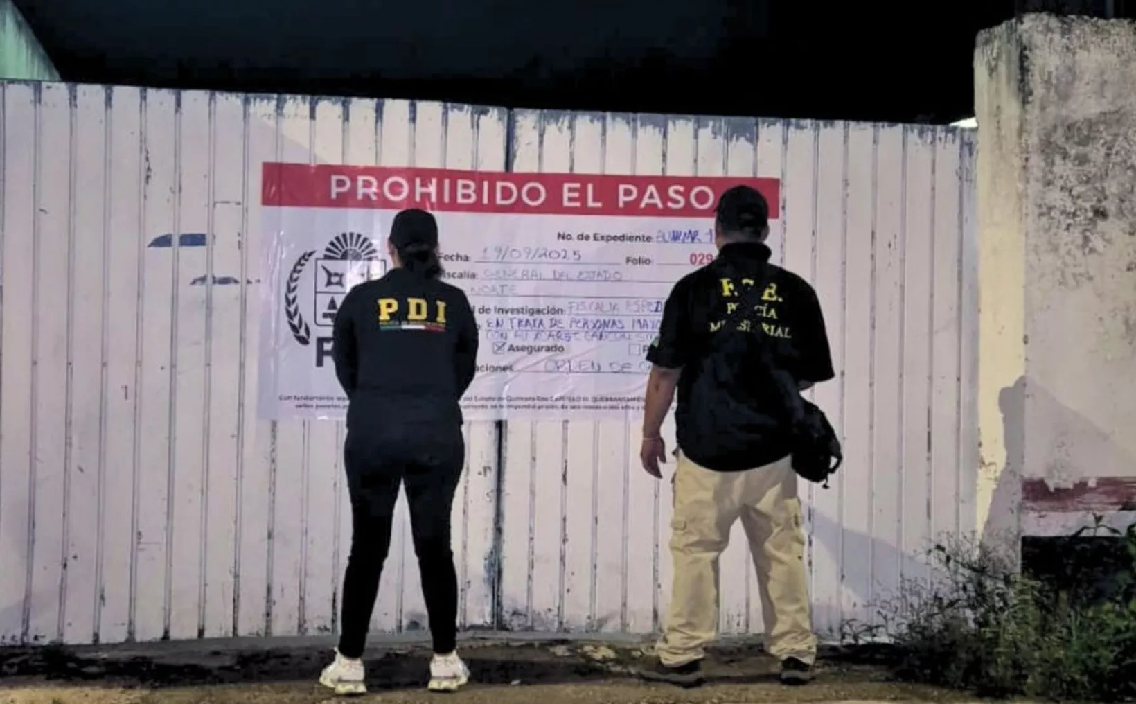 Two individuals wearing police jackets standing in front of a sign that says "Prohibido El Paso" (No Entry) at night