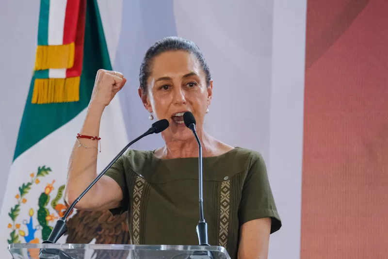 A woman speaking passionately at a podium with a microphone, raising her fist in determination, with a Mexican flag in the background.