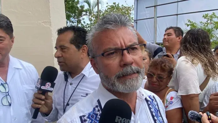 A group of people at a public event with a man in the foreground speaking to reporters and others surrounding him. He has glasses and a beard, wearing a white shirt with decorative patterns.