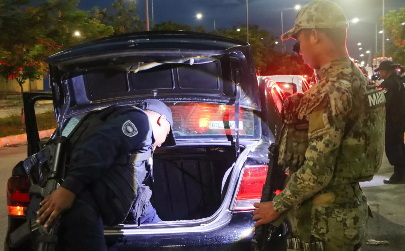 Law enforcement officers inspecting a car trunk at night with visible police presence in the background.$# CAPTION