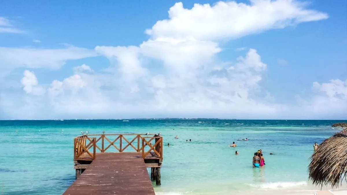 A scenic view of a wooden pier extending into a calm turquoise sea with people swimming and enjoying the beach on a sunny day under blue skies and fluffy clouds.$# CAPTION