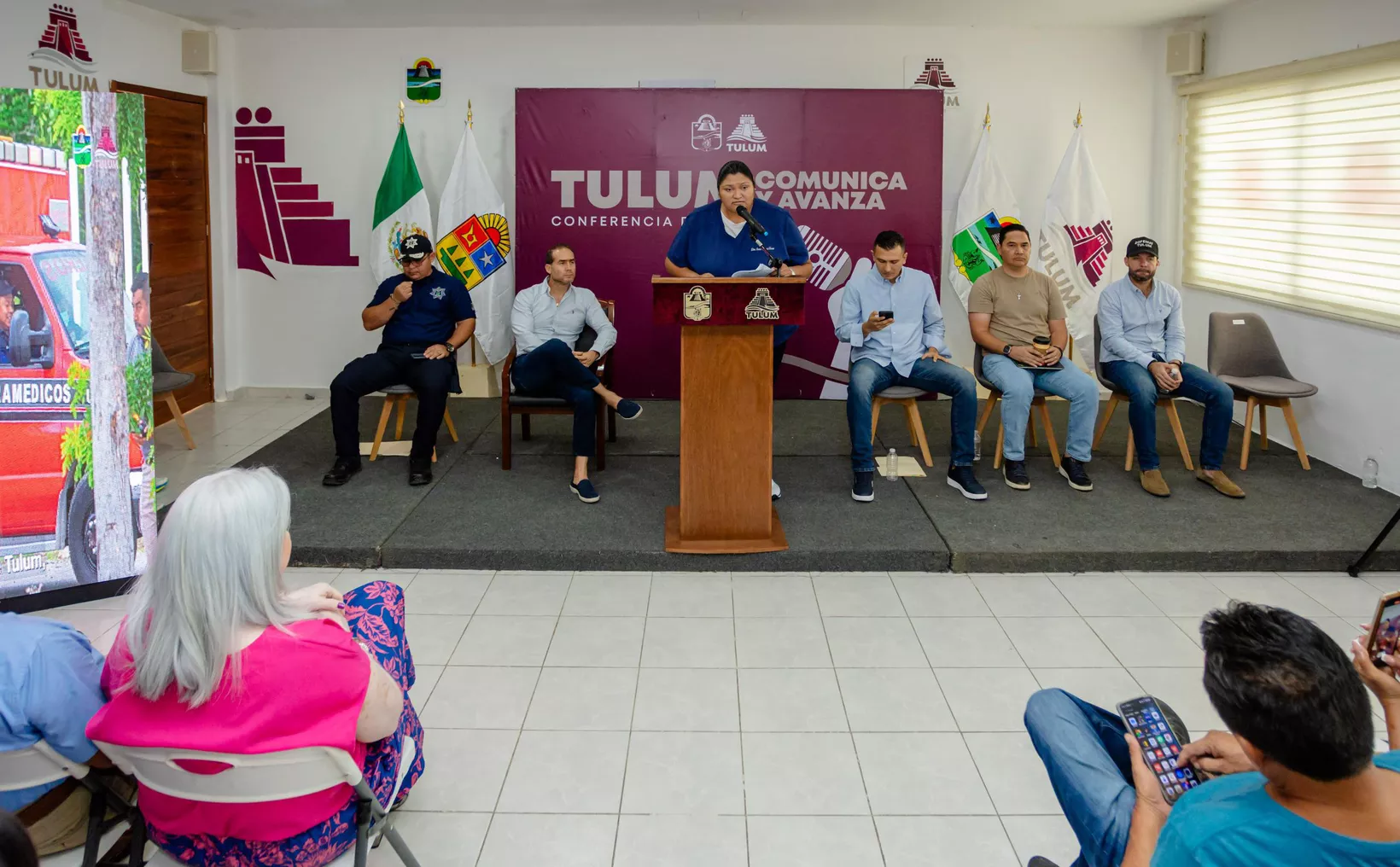 A community conference in Tulum with officials speaking at a podium, while audience members listen and engage with their devices.$# CAPTION