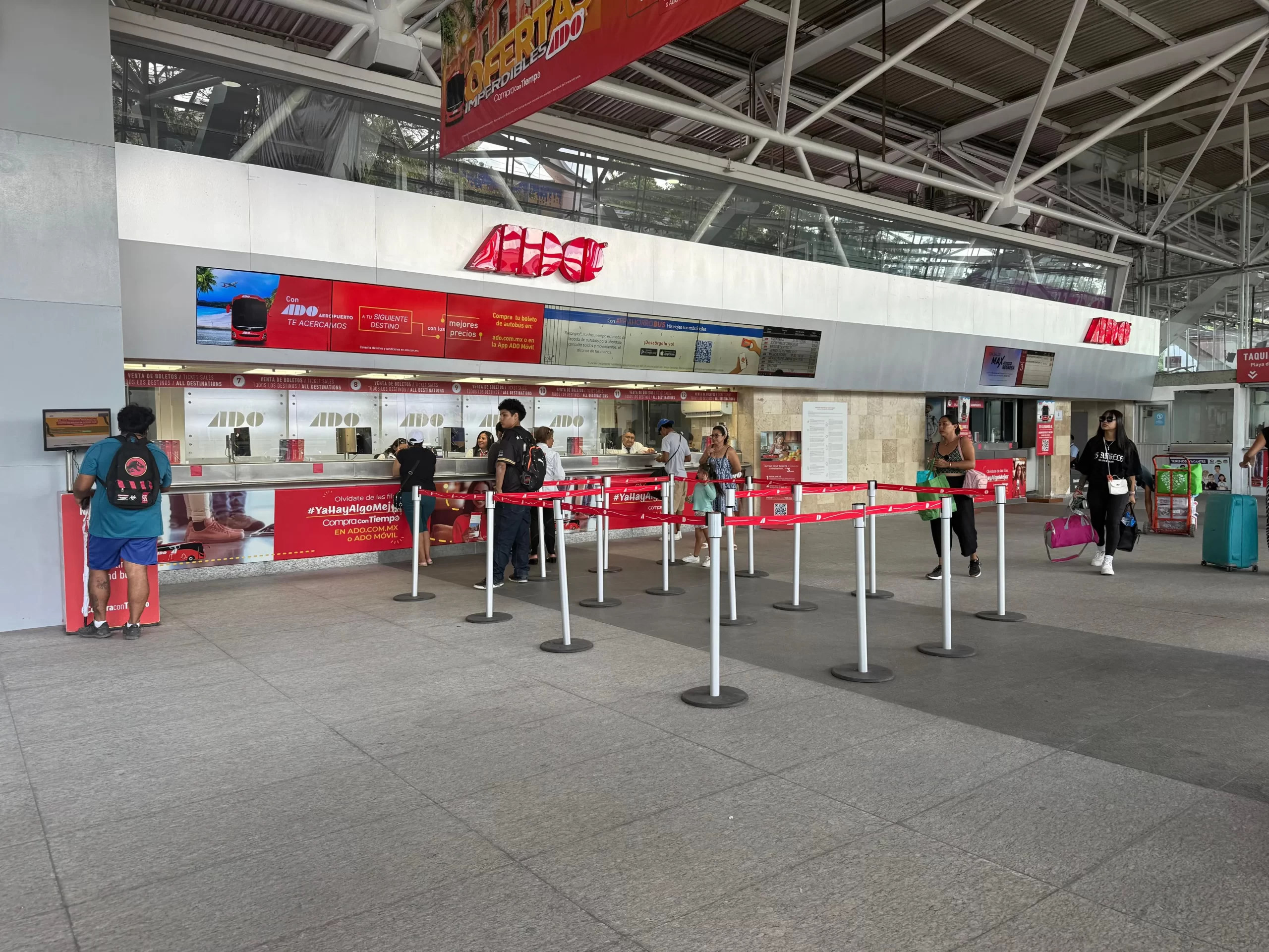 A transport terminal with ticket counters and passengers waiting in line. Some people are carrying luggage while others are at the counters.$#$ CAPTION