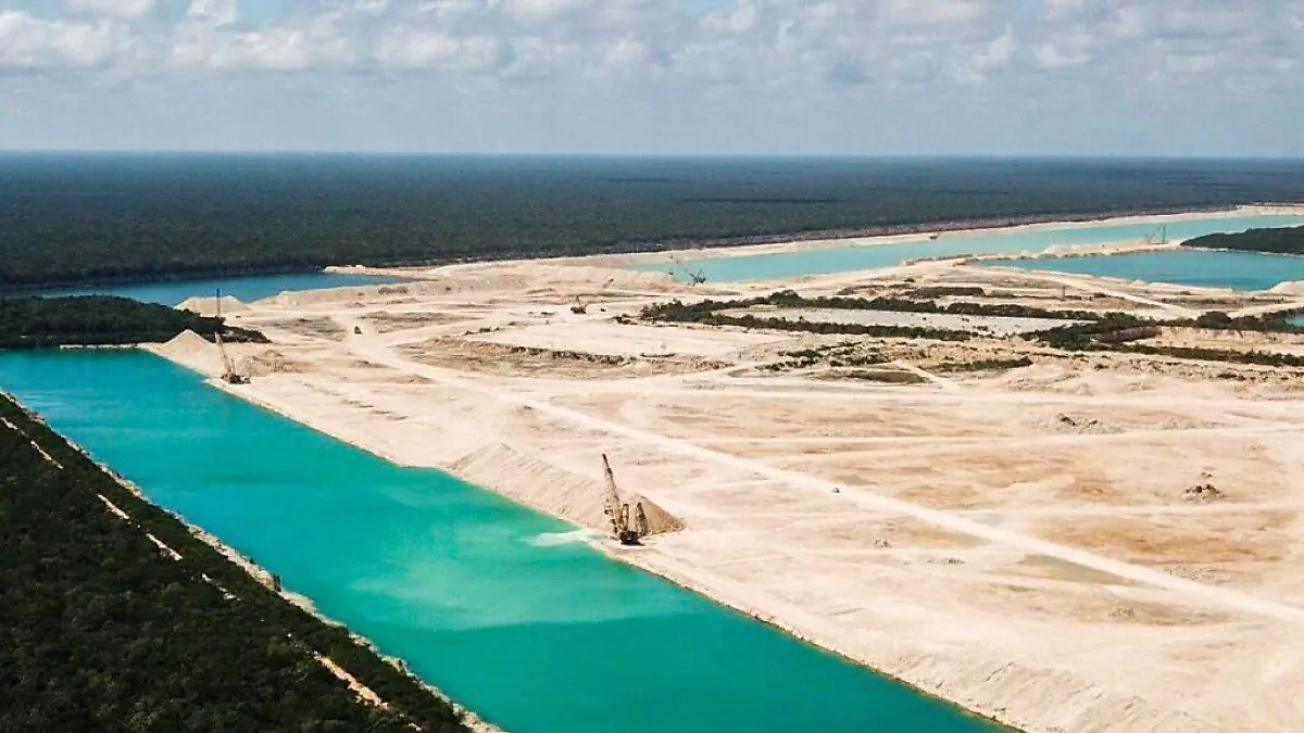 Aerial view of a landscape featuring a turquoise waterway and industrial excavation sites surrounded by greenery and scattered clouds in the sky.$#$ CAPTION