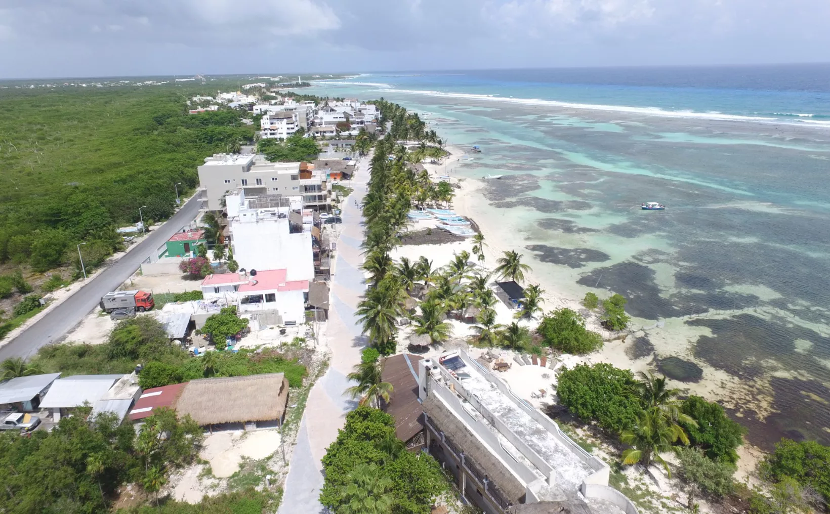 Aerial view of a coastal area featuring a sandy beach, palm trees, and buildings along the shore with turquoise waters in the background.