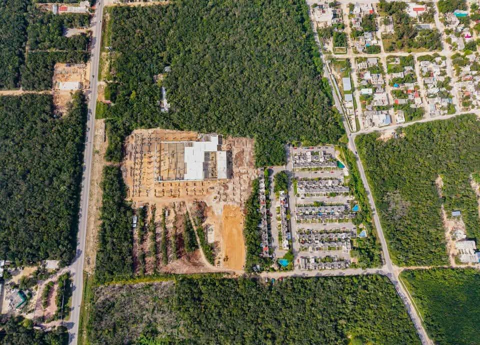 Aerial view showing a construction site alongside residential areas and a road, surrounded by greenery.$# CAPTION