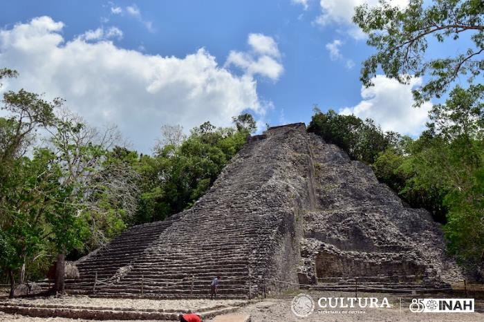 A view of an ancient Maya pyramid surrounded by lush greenery and under a partly cloudy sky.$#$ CAPTION
