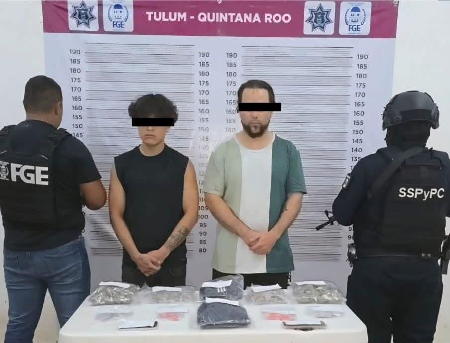 Two individuals suspected of involvement in criminal activity, flanked by law enforcement officers, with evidence displayed on a table in Tulum, Quintana Roo.$# CAPTION