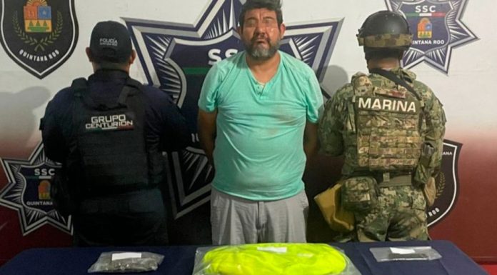 A man in a blue shirt is handcuffed between two uniformed officers, with items on a table in front of them.$# CAPTION