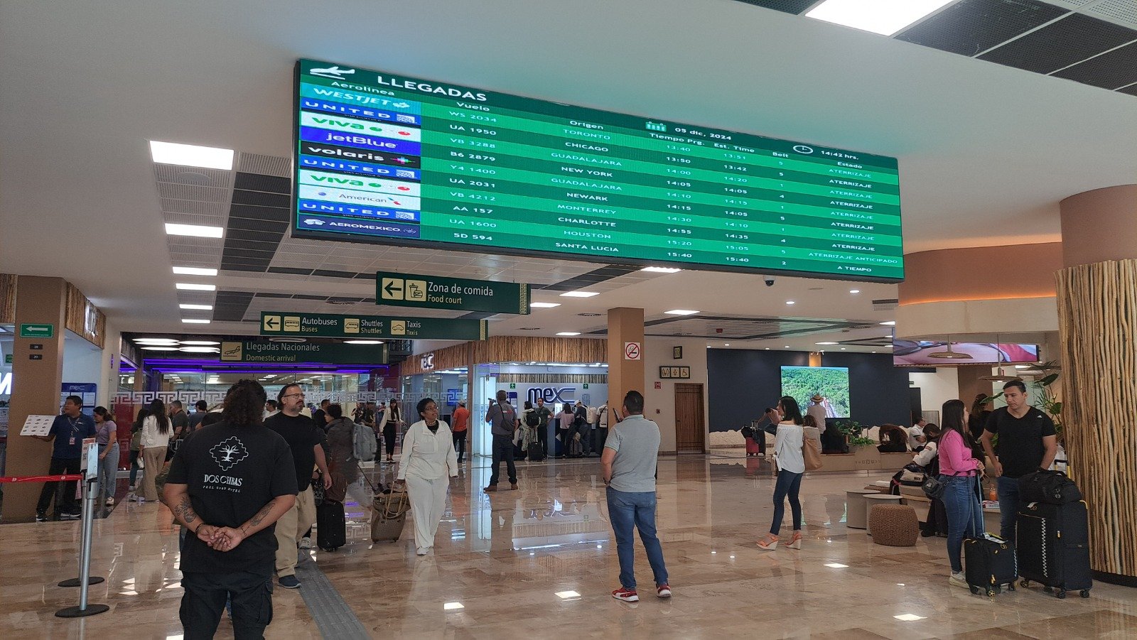 A bustling airport arrival hall with passengers and flight information screens displaying arrival details in both Spanish and English.$# CAPTION