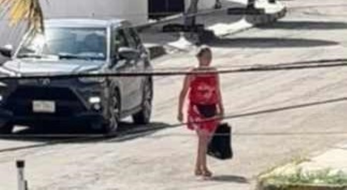 A person wearing a red dress walking on the street with a black bag, alongside a parked black SUV.