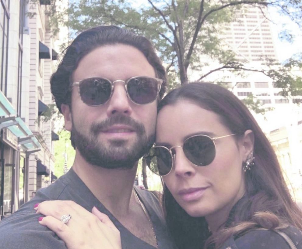 A couple poses for a close-up selfie, both wearing sunglasses and smiling, in an urban setting with street trees in the background. The woman is showing off a large engagement ring.