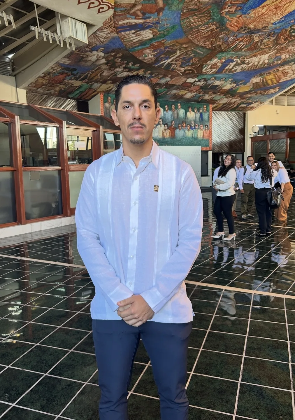 A man in a white traditional shirt and blue pants stands confidently in a spacious hall with a mural on the ceiling and people in the background.$# CAPTION