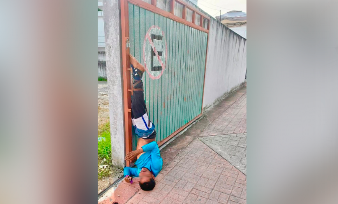 A boy hanging upside down from a gate, trying to maneuver through a narrow opening while holding a small item in his hands. He is wearing a blue shirt and black pants. The gate has a "No Parking" sign painted on it.
